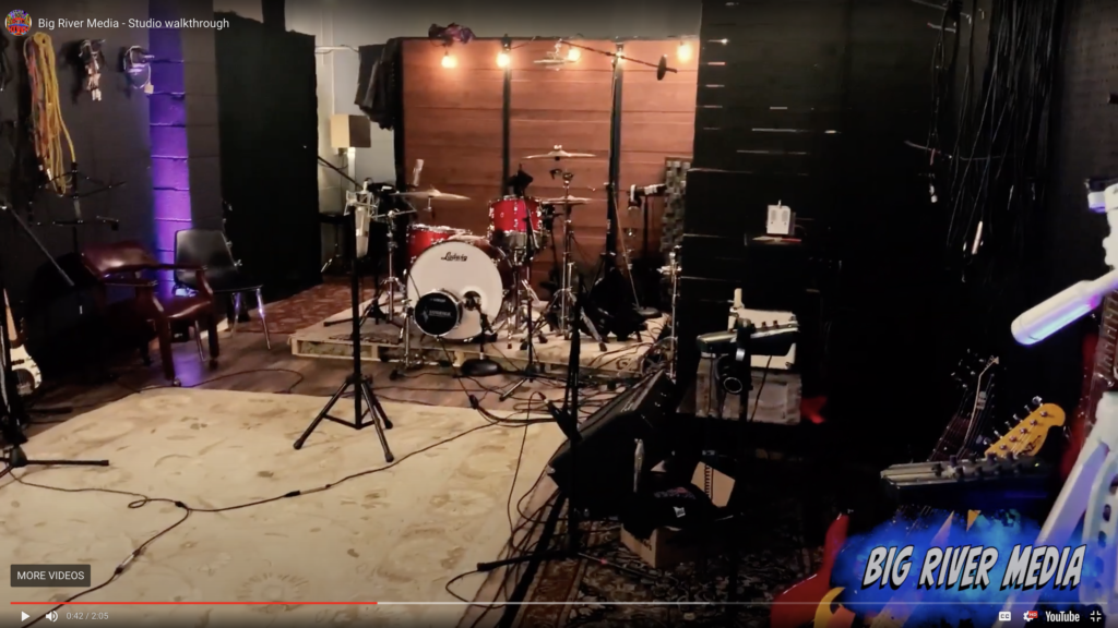 A drum kit and multiple mic stands are set up in a room full of audio recording gear.